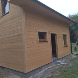 Piotr łozowski alt- drew - Surowy, drewniany dom szkieletowy z widocznymi detalami konstrukcyjnymi, małymi oknami i betonową podmurówką, na tle zieleni ogrodu.