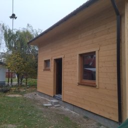 Piotr łozowski alt- drew - Surowy drewniany budynek z oknami i drzwiami, z czarną rynną spustową, w trakcie budowy, z widocznymi bloczkami betonowymi przed wejściem i elementami wyposażenia na trawniku.