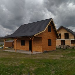 Piotr łozowski alt- drew - Nowoczesny, parterowy budynek z drewna z ciemnym dachem na tle pochmurnego nieba, widoczny w tle budynek w stanie surowym.