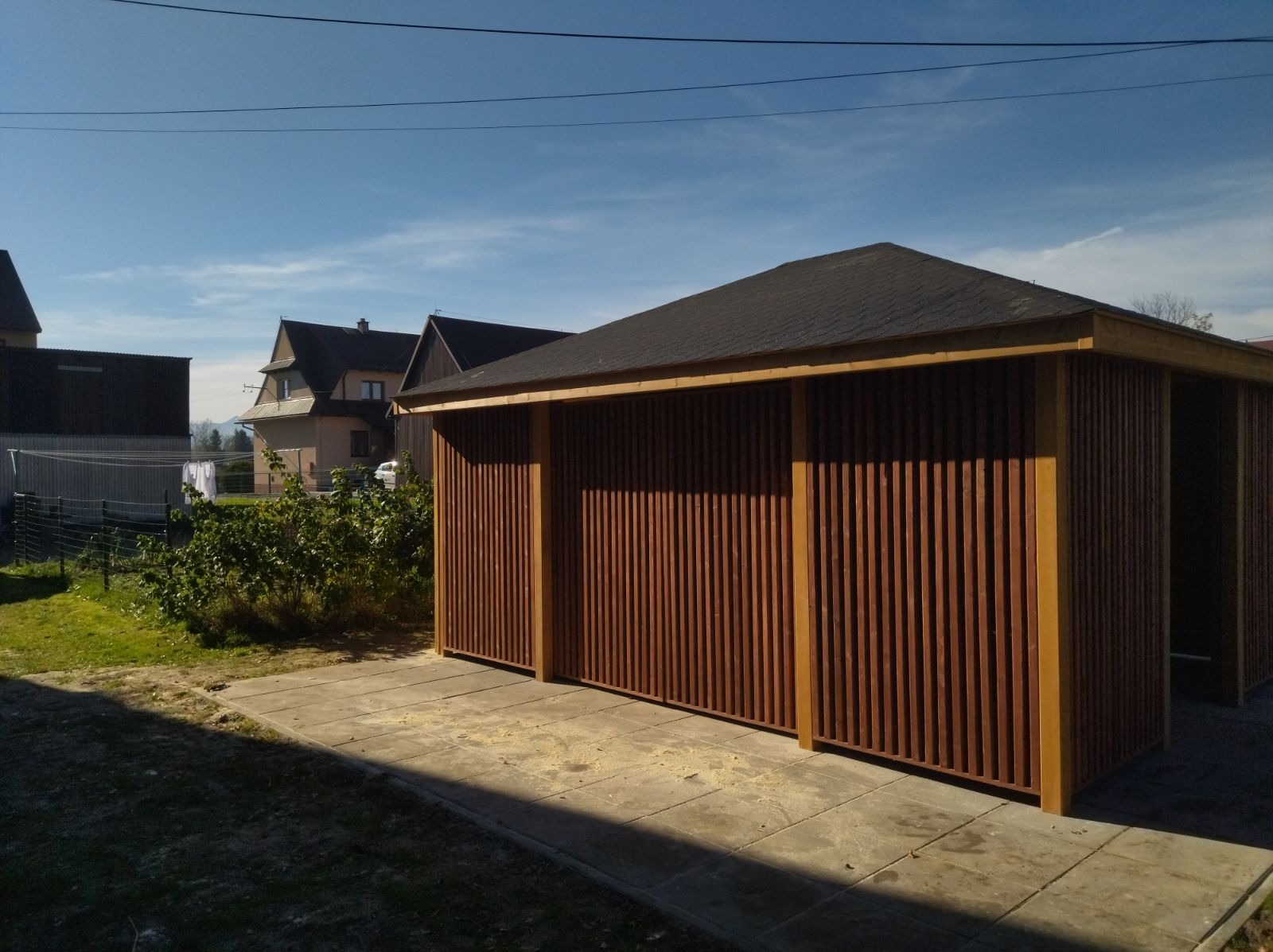 Nowoczesna konstrukcja z drewnianych listew, prawdopodobnie zadaszenie lub wiata, z czarnym dachem, na betonowej posadzce, w tle domy mieszkalne i błękitne niebo.