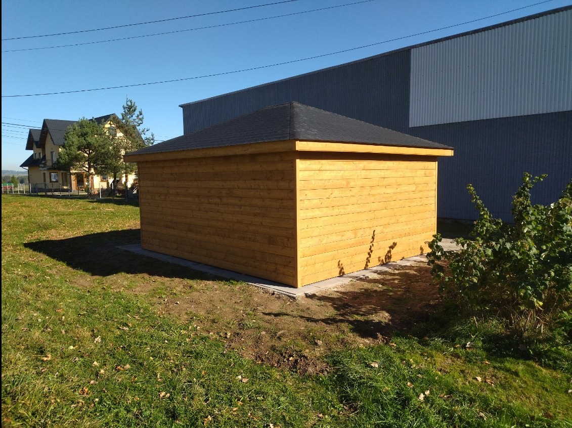 Niewielki, świeżo zaimpregnowany, drewniany budynek z czarnym, spadzistym dachem, ustawiony na betonowej podstawie na trawniku, z budynkiem gospodarczym w tle.