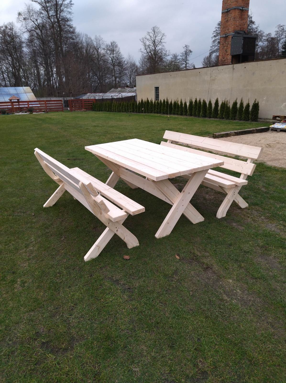 Surowy, jasny drewniany zestaw piknikowy z ławkami w kształcie litery X, ustawiony na trawniku przed budynkiem z ceglanym kominem i rzędem tuj.