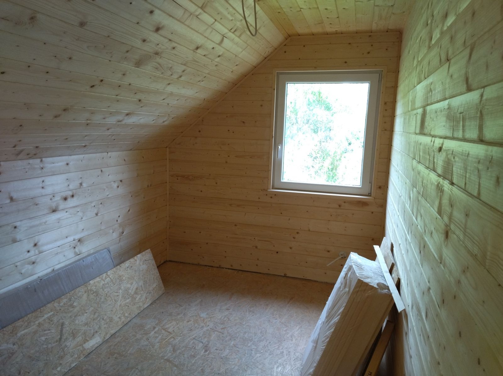 Poddasze wykończone boazerią w jasnym odcieniu drewna, okno z widokiem na zieleń, na podłodze płyty OSB i materiały budowlane w folii, ukośny sufit.