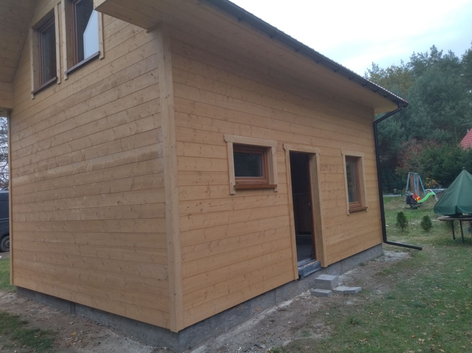 Surowy, drewniany dom szkieletowy z widocznymi detalami konstrukcyjnymi, małymi oknami i betonową podmurówką, na tle zieleni ogrodu.