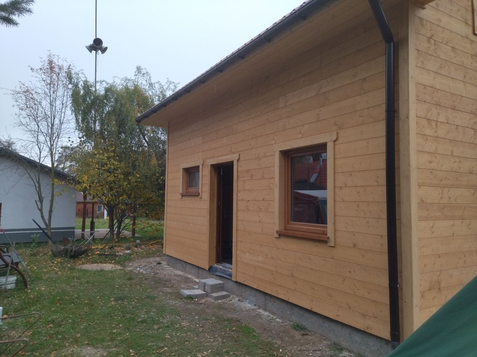 Surowy drewniany budynek z oknami i drzwiami, z czarną rynną spustową, w trakcie budowy, z widocznymi bloczkami betonowymi przed wejściem i elementami wyposażenia na trawniku.