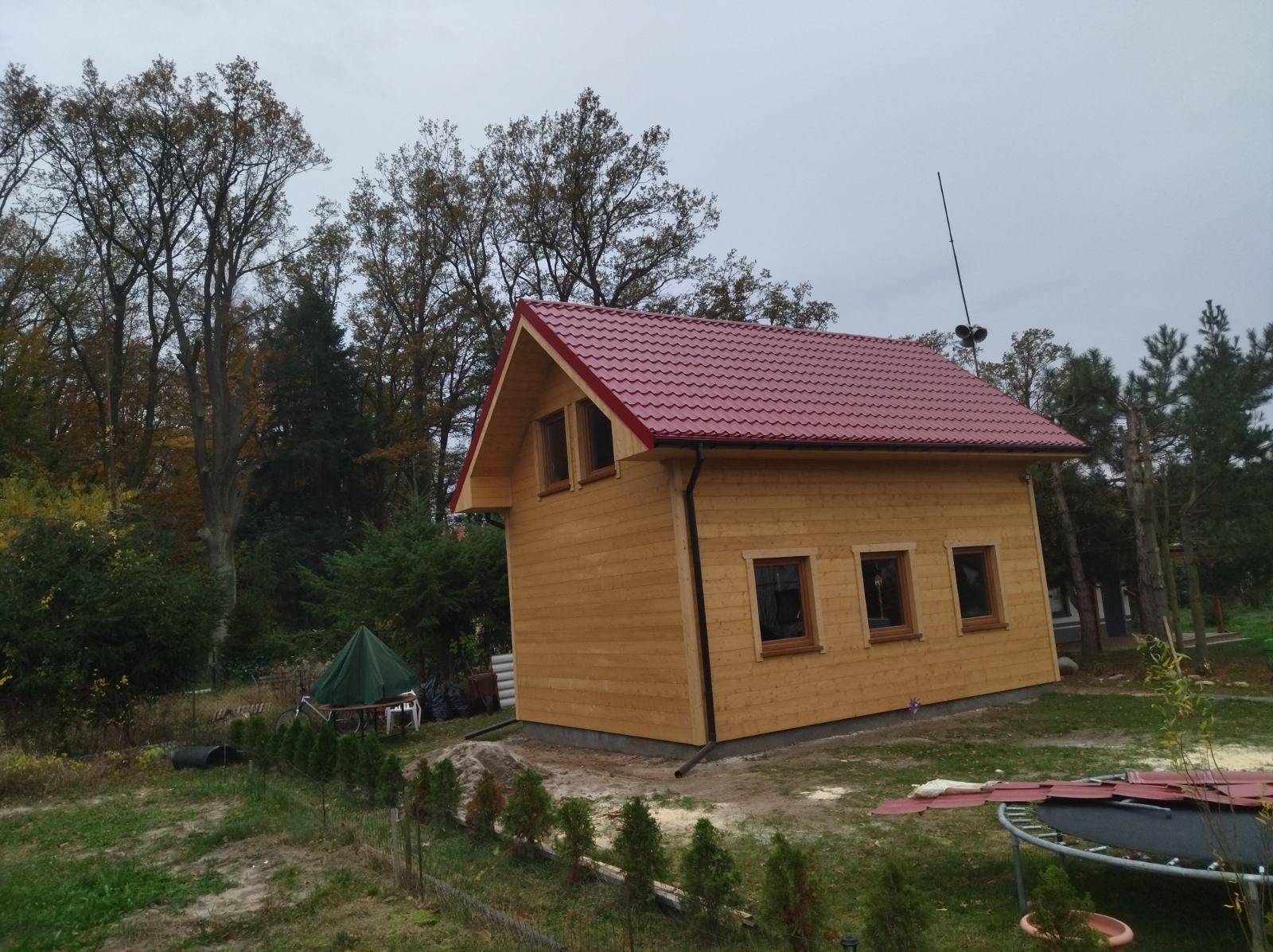 Nowo wybudowany drewniany domek letniskowy z czerwonym dachem na działce z drzewami, widok z boku, z widocznymi elementami wykończeniowymi.