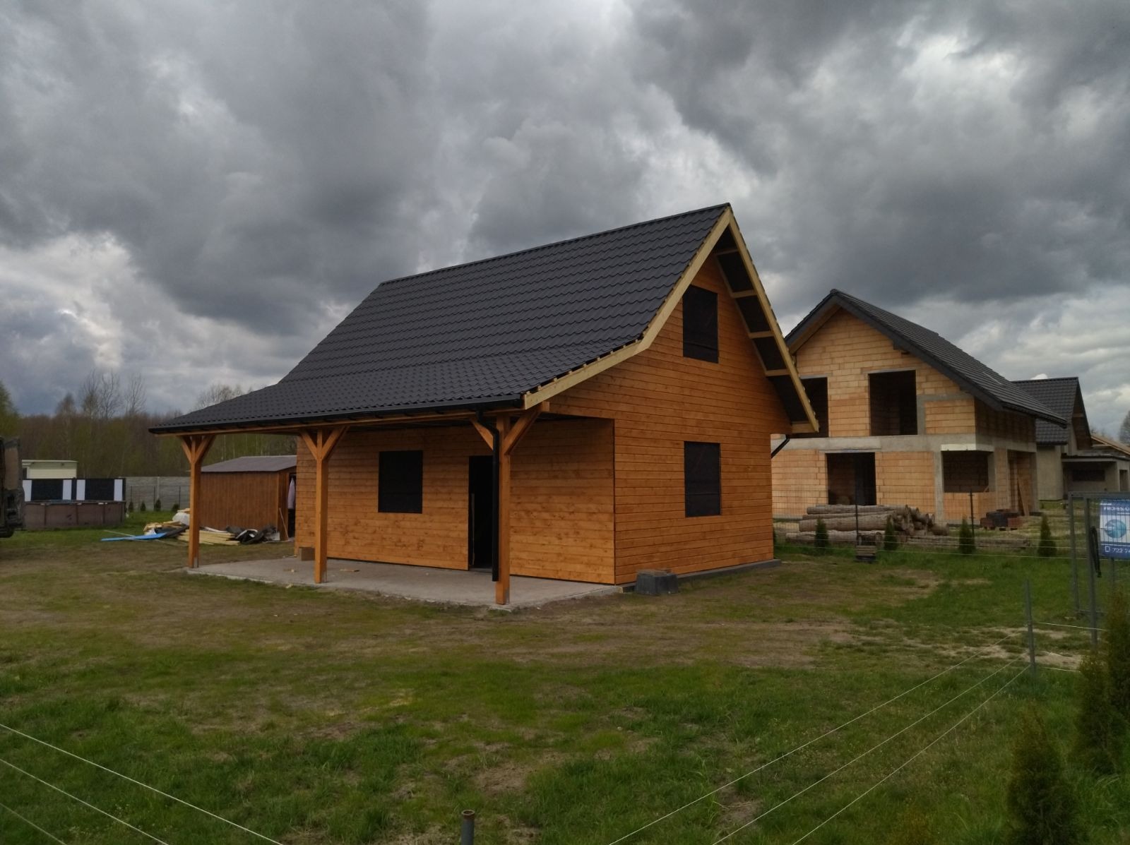 Nowoczesny, parterowy budynek z drewna z ciemnym dachem na tle pochmurnego nieba, widoczny w tle budynek w stanie surowym.