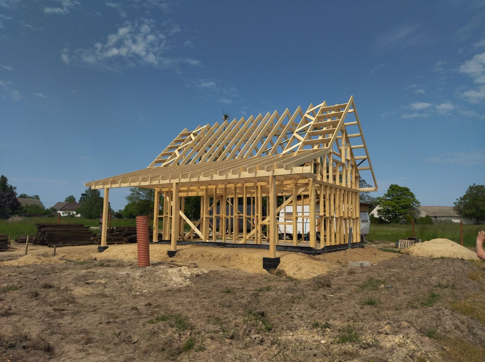 Surowa konstrukcja drewniana domu w trakcie budowy, widoczne elementy szkieletu, dach w budowie, rury wentylacyjne w pobliżu, ziemia wokół.
