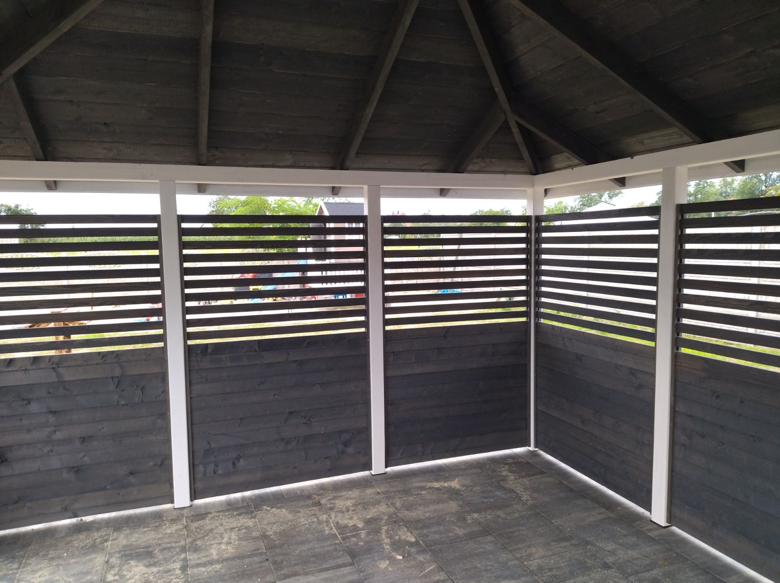 Wnętrze zadaszonej konstrukcji z drewnianymi panelami z poziomymi szczelinami, pomalowanymi na ciemny kolor. Widoczna drewniana konstrukcja dachu i szara kostka brukowa na podłodze. W tle zieleń...