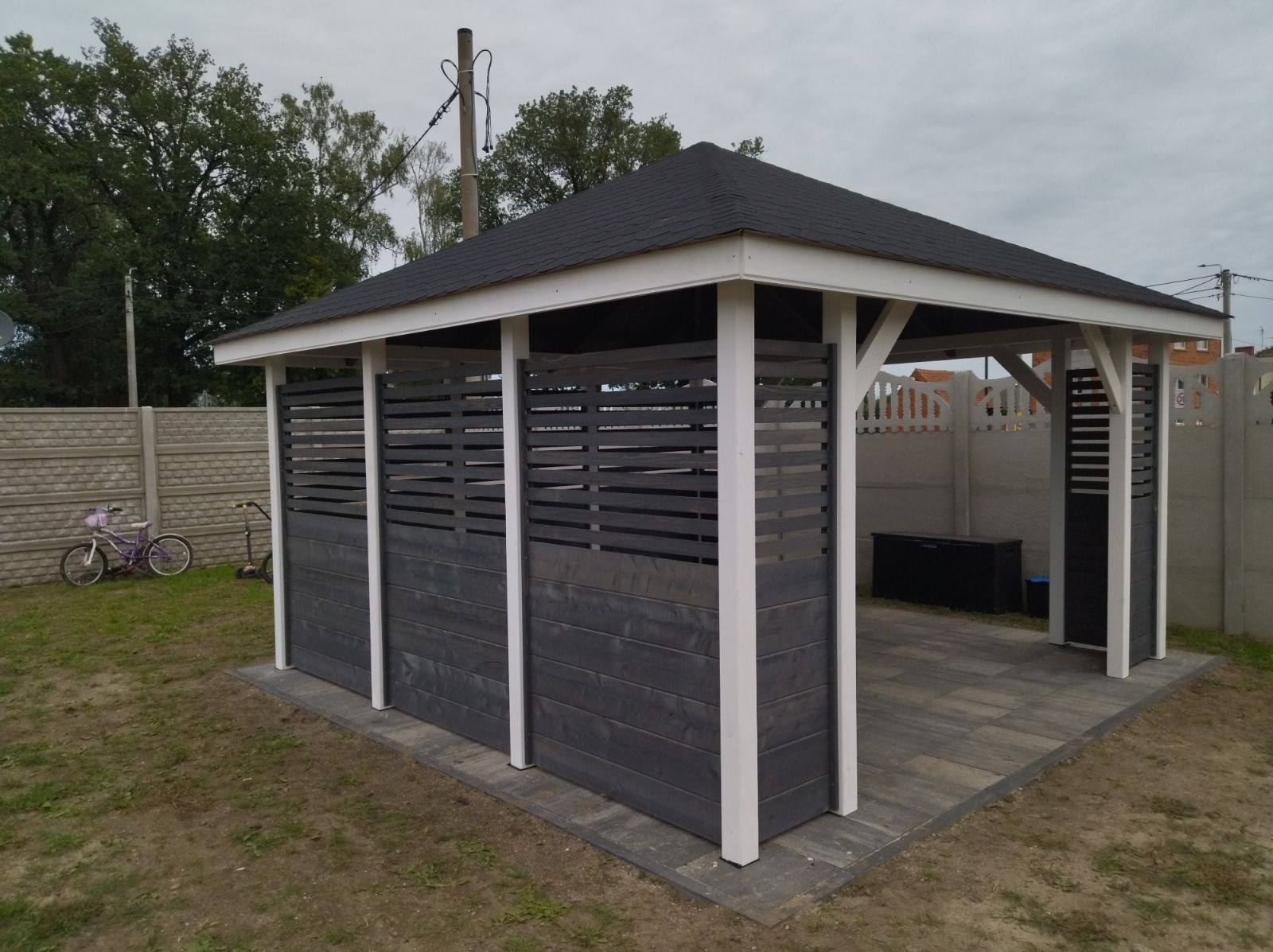 Nowoczesna altana ogrodowa z szarymi drewnianymi panelami i białymi słupkami, pokryta czarnym dachem z gontu bitumicznego, na tle betonowego ogrodzenia i pochmurnego nieba.