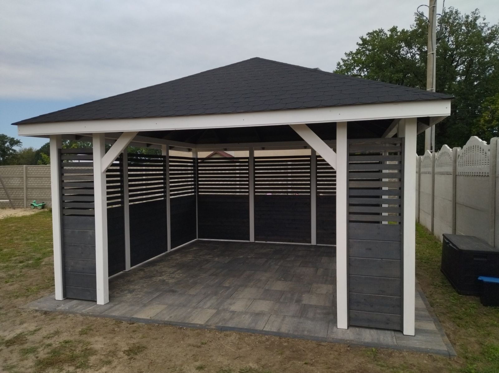 Nowoczesna altana ogrodowa z czarnym dachem z gontu bitumicznego, szarymi panelami bocznymi z poziomych desek i białymi słupami, wyłożona szarą kostką brukową, na tle zieleni i betonowego ogrodzenia.