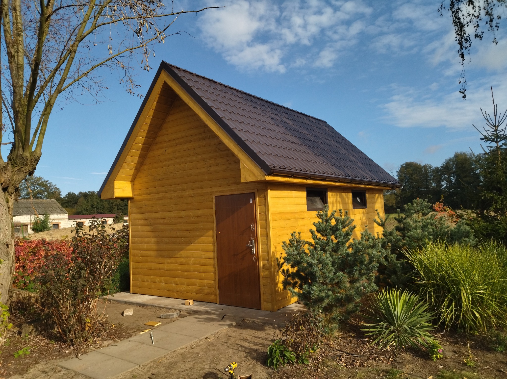 Żółty, drewniany domek narzędziowy z brązowym dachem dwuspadowym, widoczny z zewnątrz, otoczony zielenią i częściowo wyłożony kostką brukową. W tle budynek gospodarczy i drzewa.