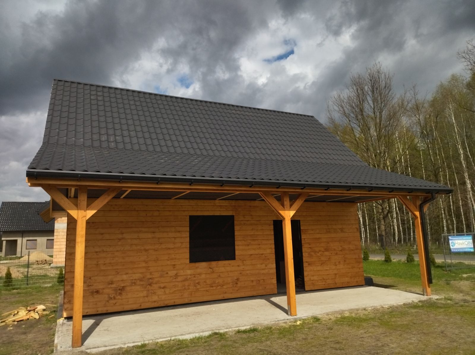 Drewniany budynek z ciemnym dachem, wsparty na drewnianych słupach, z widoczną konstrukcją dachu i fragmentem innego budynku w tle, na tle pochmurnego nieba.