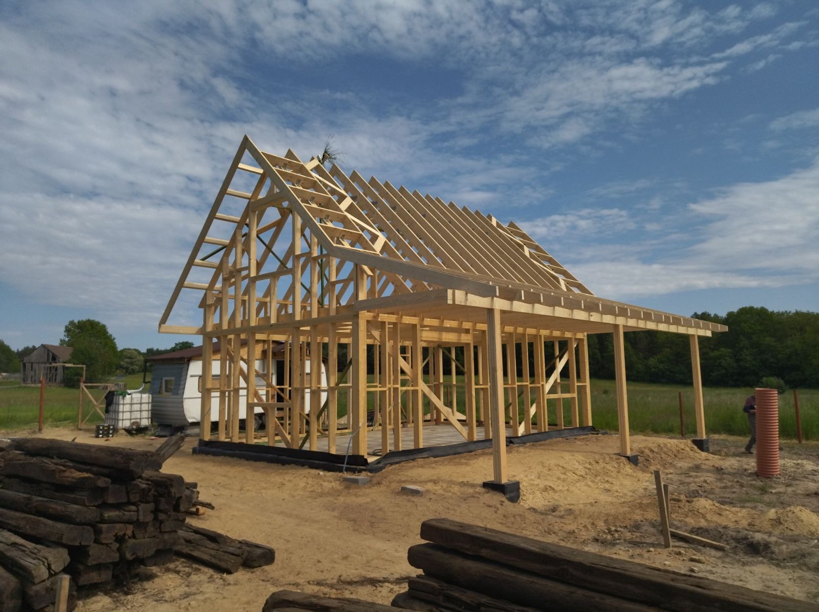 Konstrukcja szkieletowa domu w trakcie budowy, widoczne drewniane elementy nośne, dach w budowie, w tle przyczepa kempingowa i stary budynek gospodarczy, na pierwszym planie stos drewnianych bali.