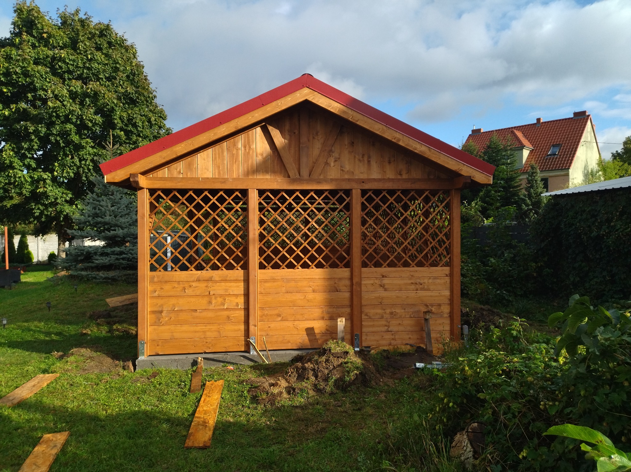 Nowo postawiona drewniana konstrukcja z czerwonym dachem, z kratowanymi panelami bocznymi, na trawniku z widocznymi śladami prac ziemnych, w tle dom z czerwoną dachówką.
