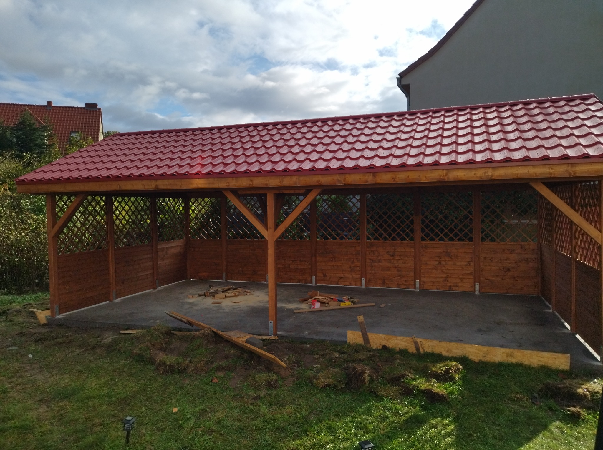 Konstrukcja zadaszona z czerwonym dachem z blachodachówki, drewnianymi słupami i ażurowymi ściankami, w trakcie budowy, widoczne narzędzia i resztki materiałów budowlanych.