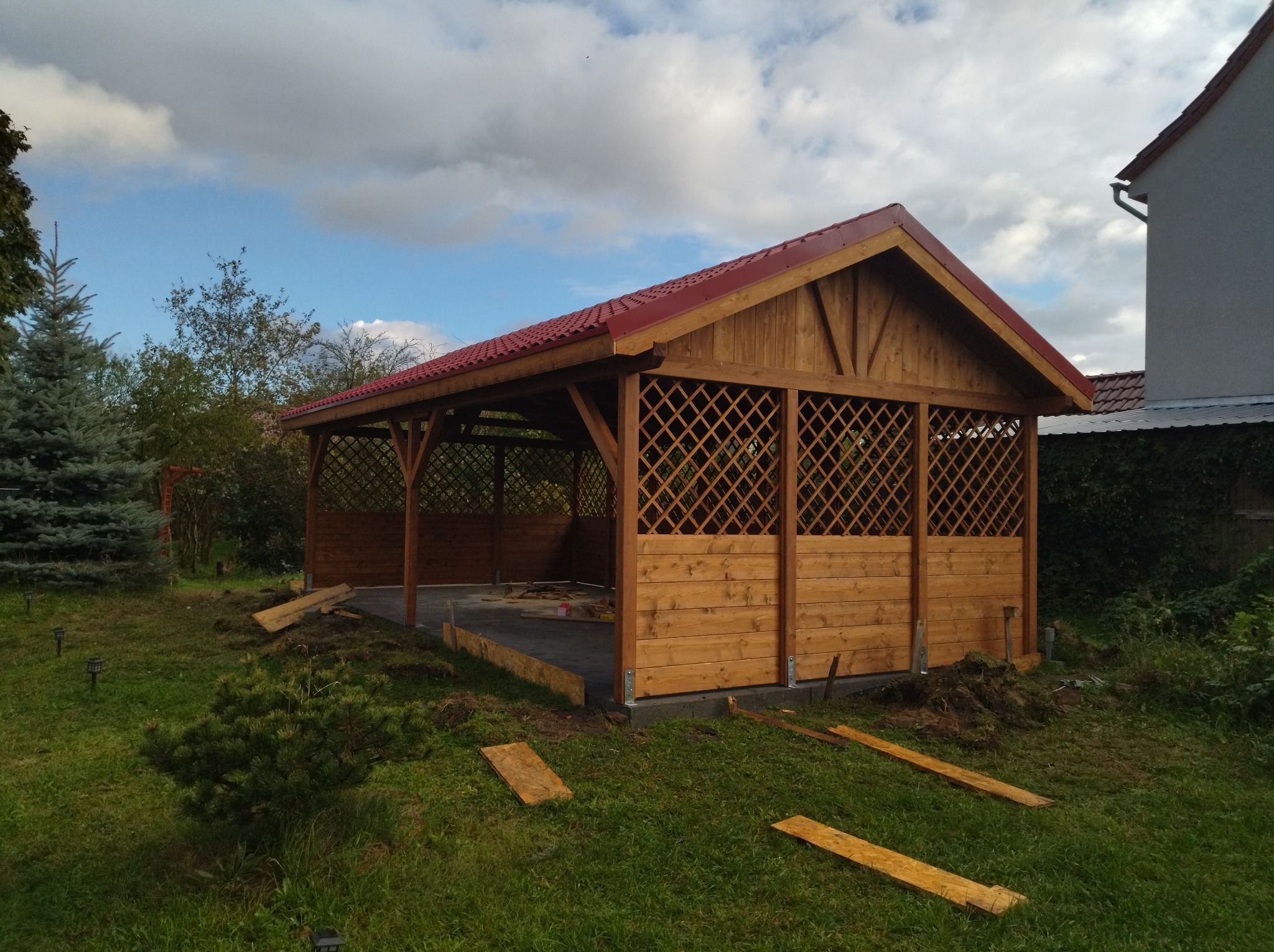 Drewniana altana z czerwoną dachówką w trakcie budowy, widoczne elementy konstrukcyjne i przygotowany teren.