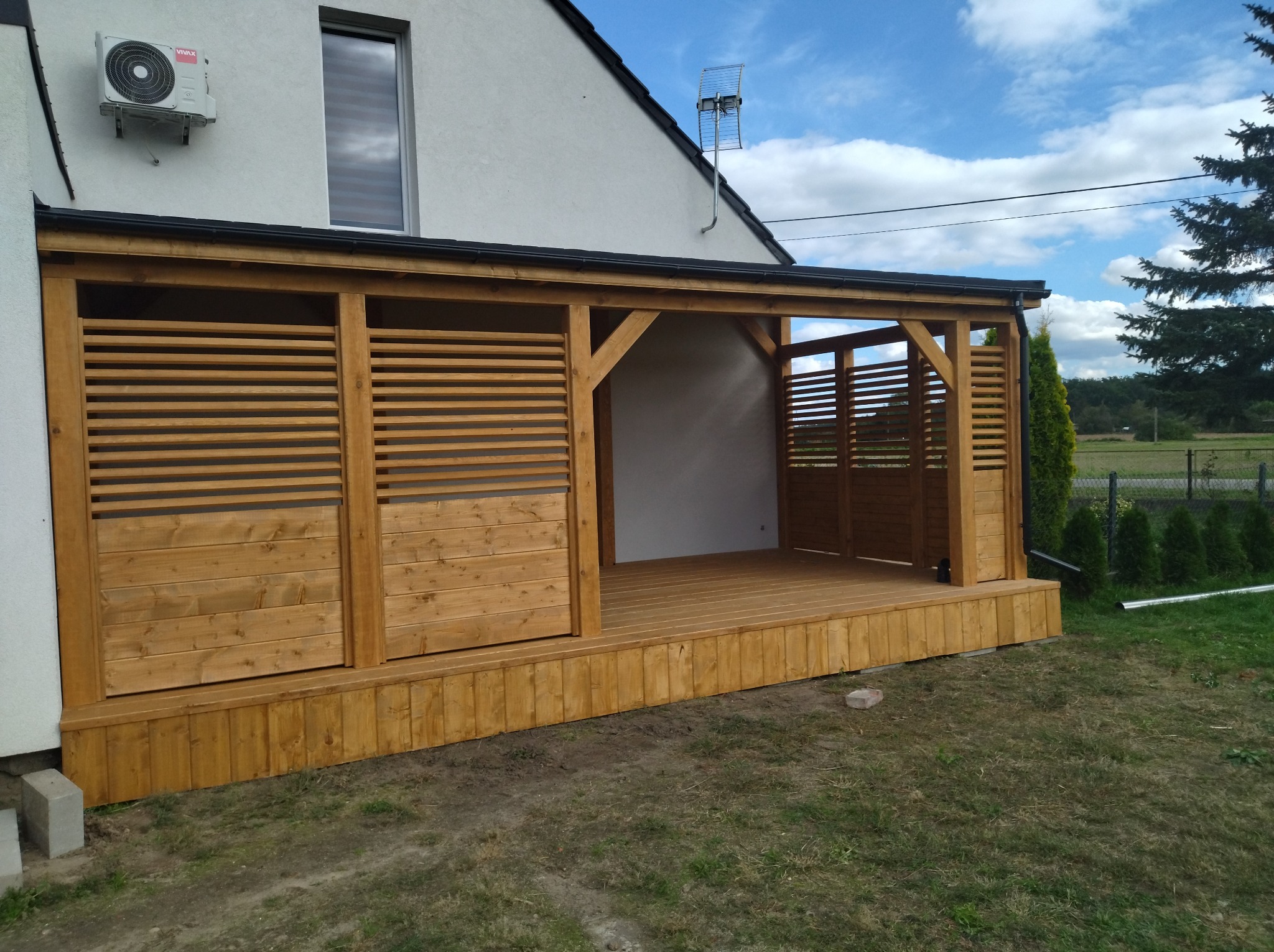 Zadaszona konstrukcja z drewna z ażurowymi ścianami w Miliczu, przylegająca do budynku mieszkalnego z klimatyzatorem na elewacji.