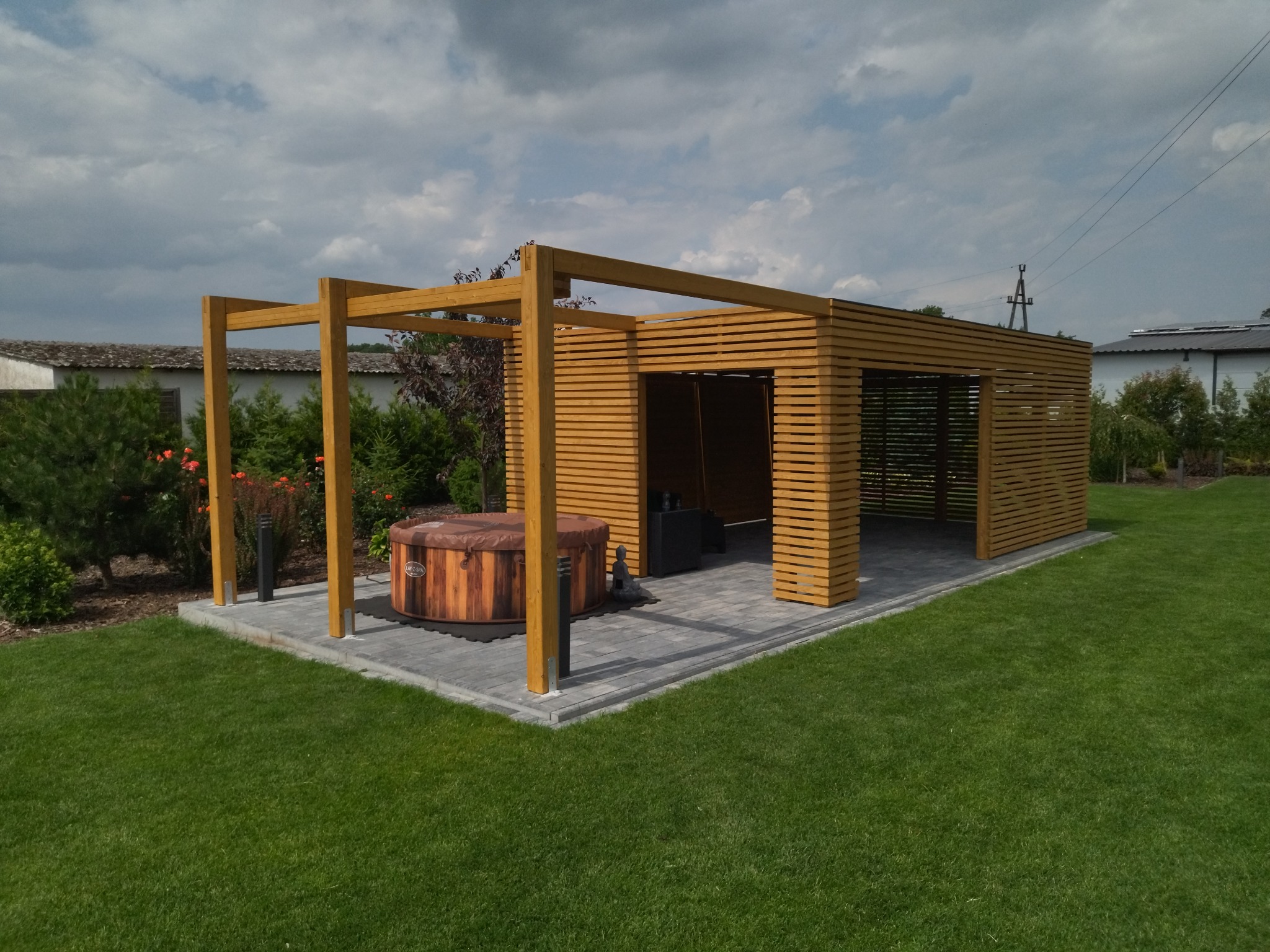 Nowoczesna, otwarta konstrukcja z drewna z poziomymi listwami, kryjąca jacuzzi z pokrywą, ustawiona na wybrukowanym tarasie, otoczona zielonym trawnikiem i roślinnością.
