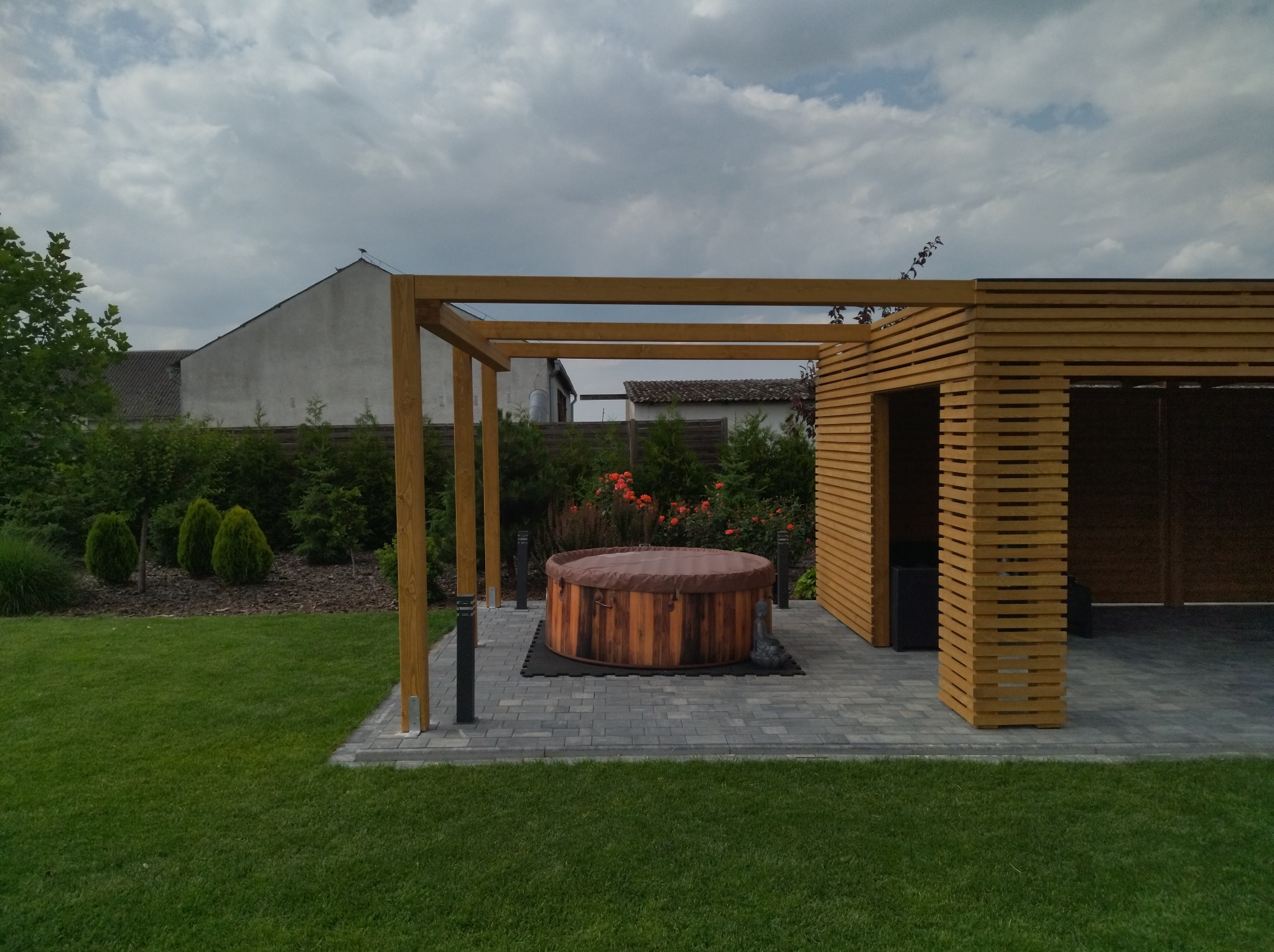 Nowoczesna drewniana konstrukcja z zadaszeniem, obok której znajduje się drewniana wanna typu jacuzzi z pokrywą, ustawiona na szarym bruku, otoczona zielonym trawnikiem i roślinnością.