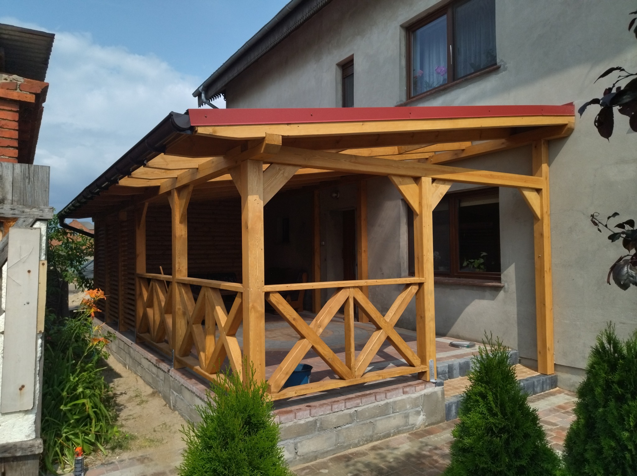 Zadaszony drewniany taras z balustradą w kształcie krzyży, przylegający do domu, z widocznymi elementami konstrukcyjnymi i czerwoną blachą na dachu, otoczony zielenią i kostką brukową.