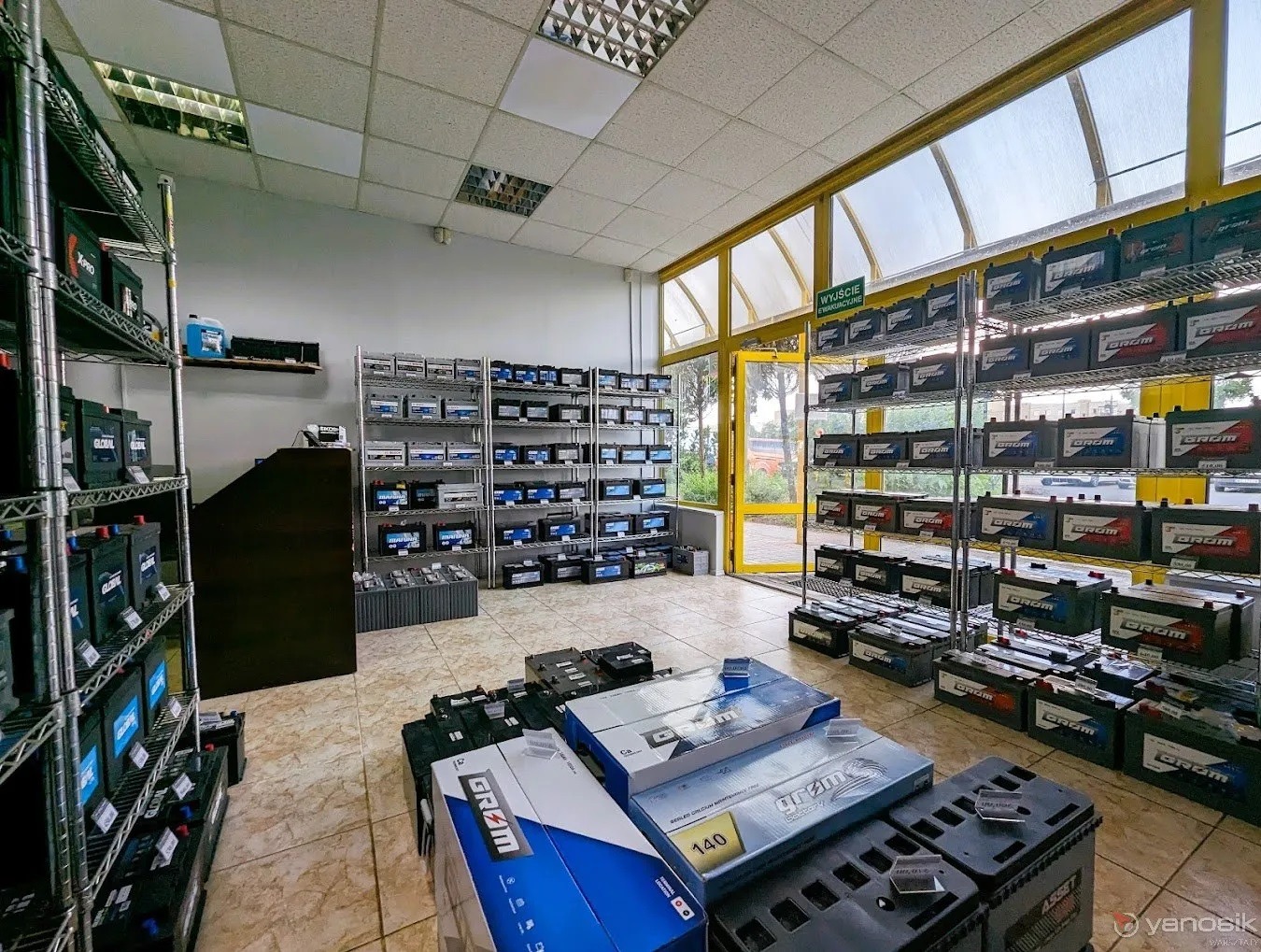 Sklep z akumulatorami samochodowymi: Rzędy półek wypełnione akumulatorami różnych marek, w tym Grom i Asset, ułożone na podłodze w opakowaniach i bez, widoczne przez przeszklone ściany wejście...