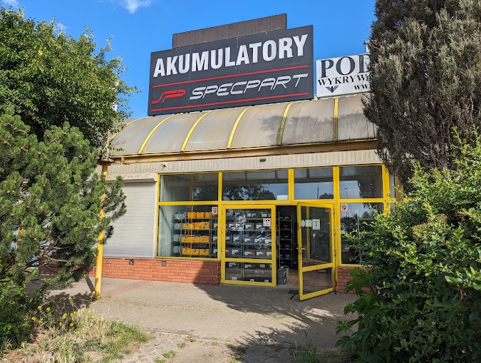 Fasada sklepu z akumulatorami samochodowymi Specpart z otwartymi drzwiami, widoczne półki z akumulatorami wewnątrz, tablica reklamowa na dachu budynku.