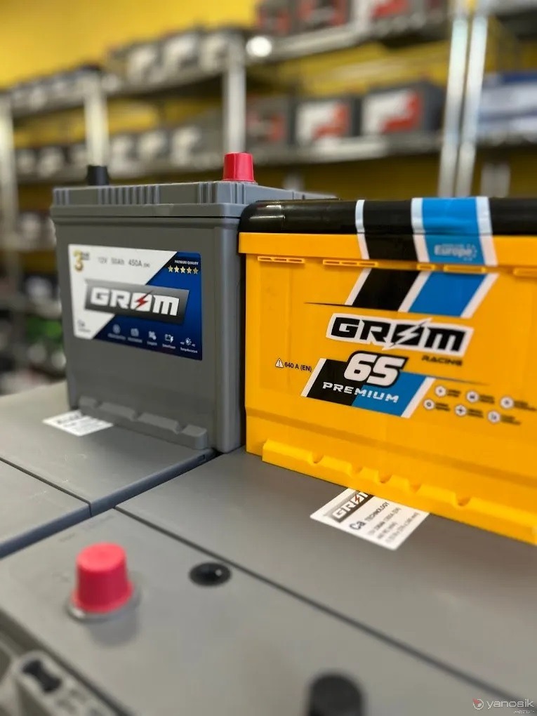 Dwa akumulatory samochodowe marki GRAM, jeden szary i jeden żółty, stojące na tle półek sklepowych. Widoczne etykiety z parametrami i logo producenta. Akumulator szary ma czerwony korek.