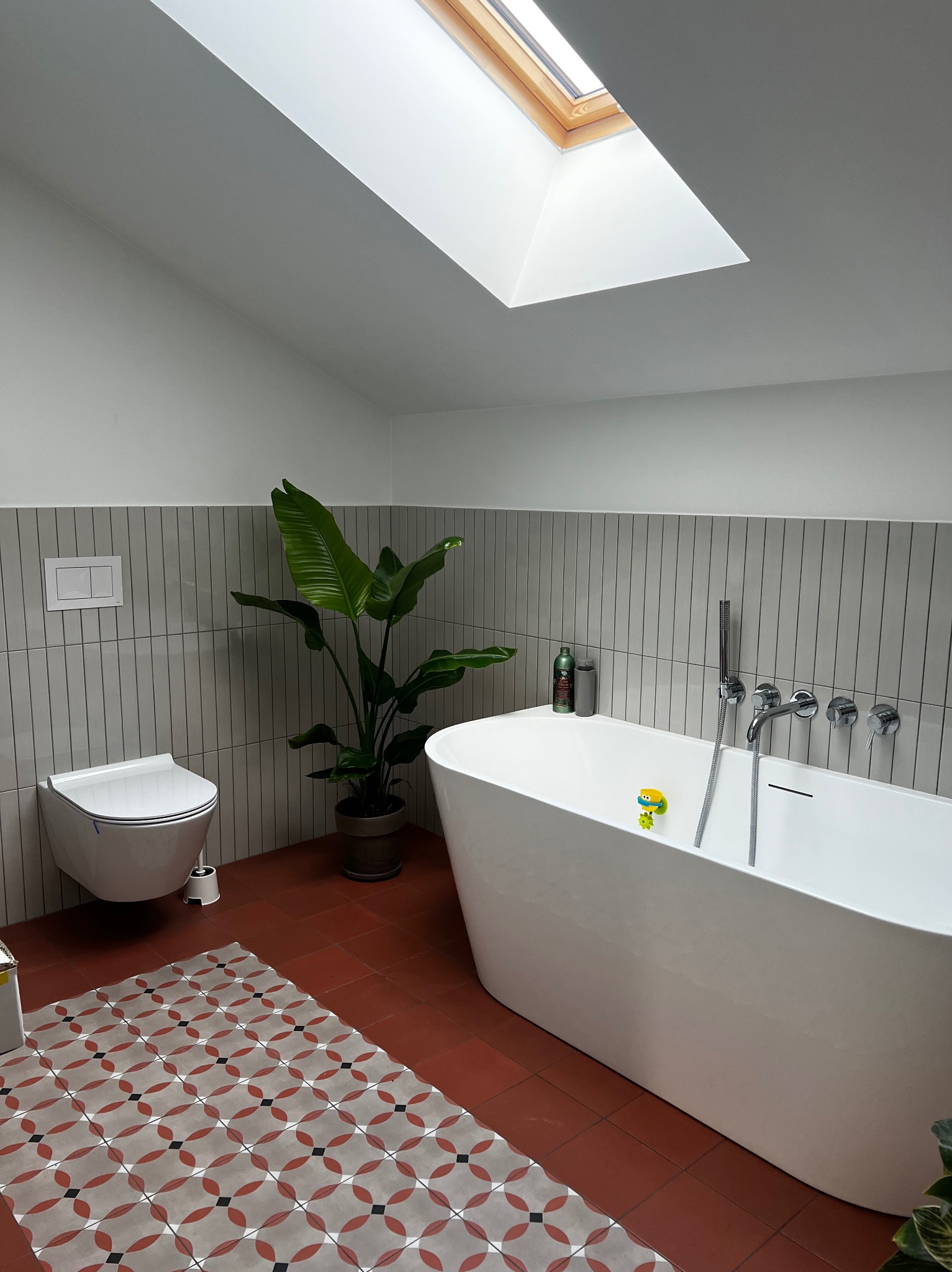 Nowoczesna łazienka z wolnostojącą wanną, terakotową podłogą w geometryczny wzór, pionowymi szarymi płytkami, rośliną doniczkową i oknem dachowym.
