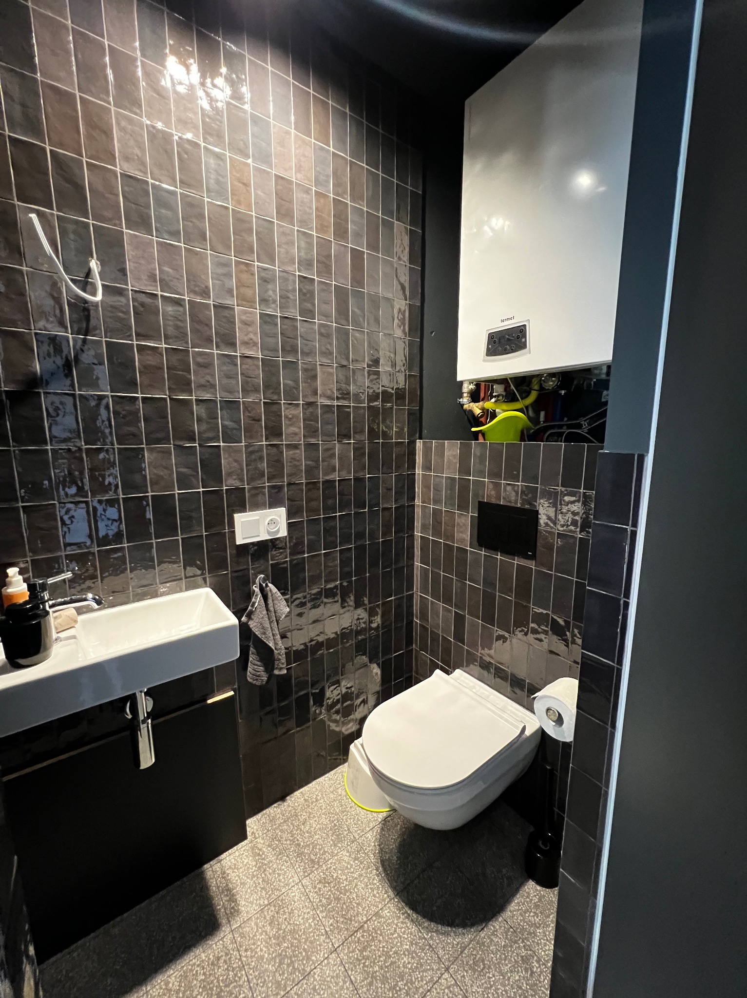 Ciemna łazienka z kwadratowymi, brązowo-czarnymi płytkami na ścianach i podłodze, wisząca umywalka z czarną szafką, toaleta z podwieszanym stelażem i wiszący piec gazowy Terma.