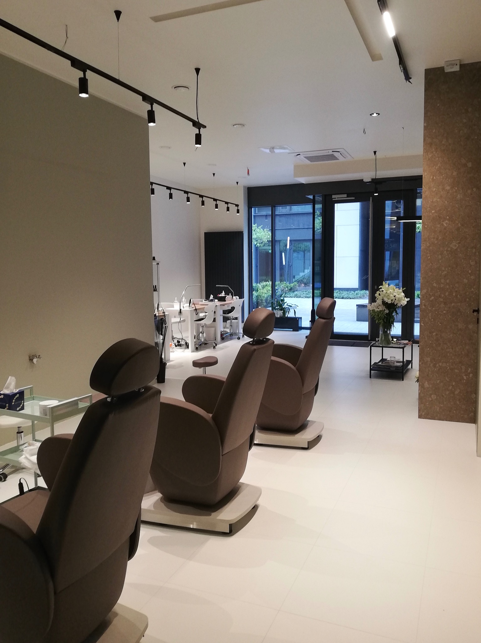 Nowoczesne wnętrze salonu kosmetycznego z trzema brązowymi fotelami i stanowiskami do manicure przy ścianie, widok na przeszklone drzwi z roślinami na zewnątrz.