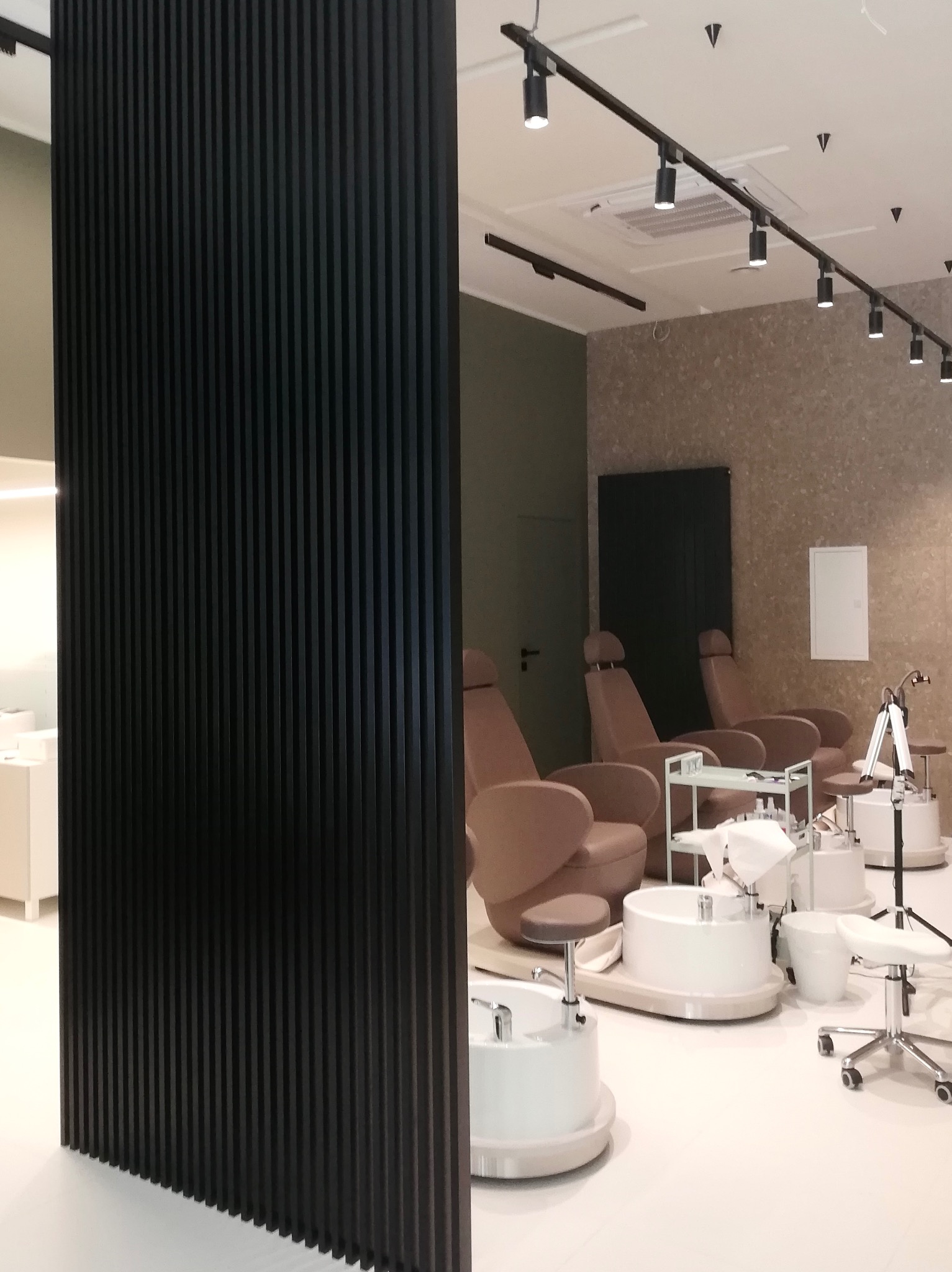Salon kosmetyczny: stanowiska do pedicure z beżowymi fotelami, białymi umywalkami i czarną, pionową ścianką działową z lameli.