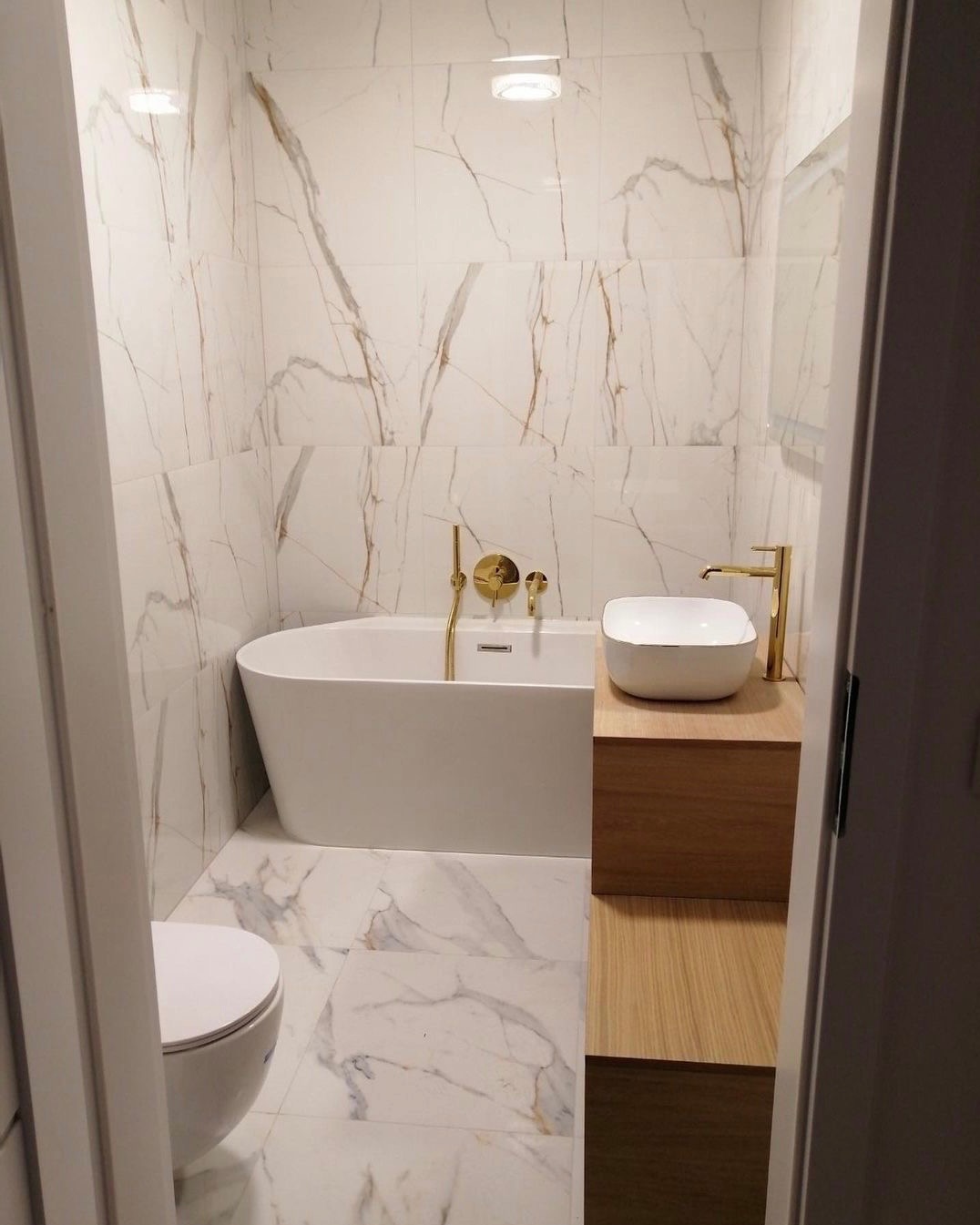 Nowoczesna łazienka z białymi płytkami imitującymi marmur, wolnostojąca wanna i umywalka nablatowa z armaturą w kolorze złotym, widok zza uchylonych drzwi.