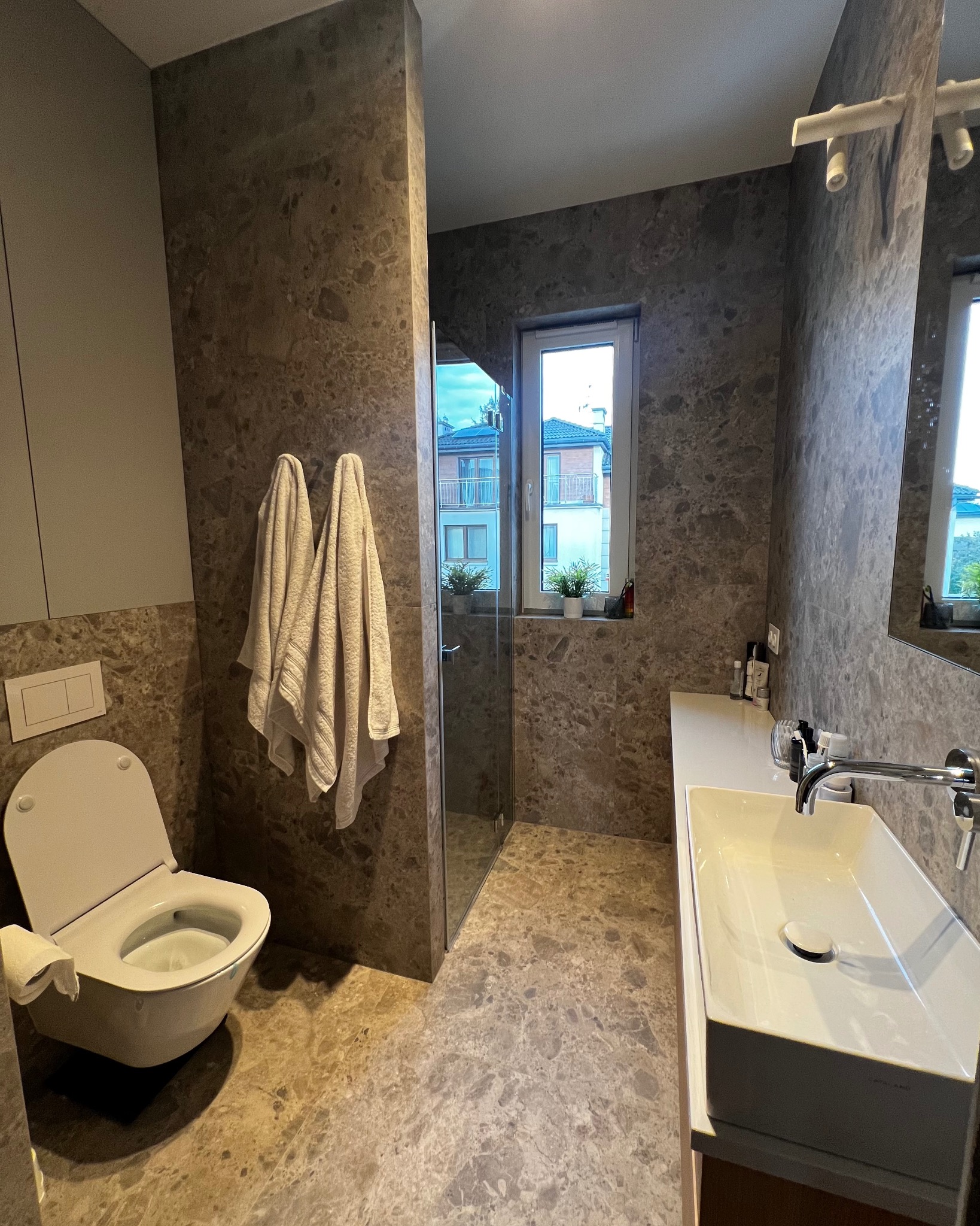 Nowoczesna łazienka z marmurowymi płytkami, toaletą z otwartą klapą, umywalką nablatową i kabiną prysznicową, widok na budynek mieszkalny przez okno.