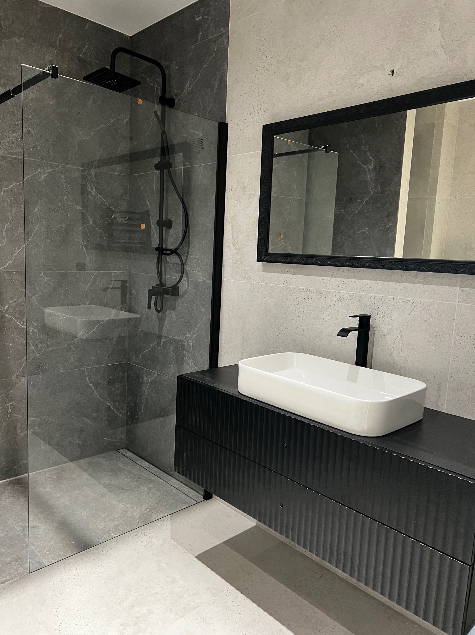 Nowoczesna łazienka z kabiną prysznicową typu walk-in z czarną armaturą, białą prostokątną umywalką nablatową na czarnej szafce z ryflowanymi frontami i lustrem w ozdobnej czarnej ramie.