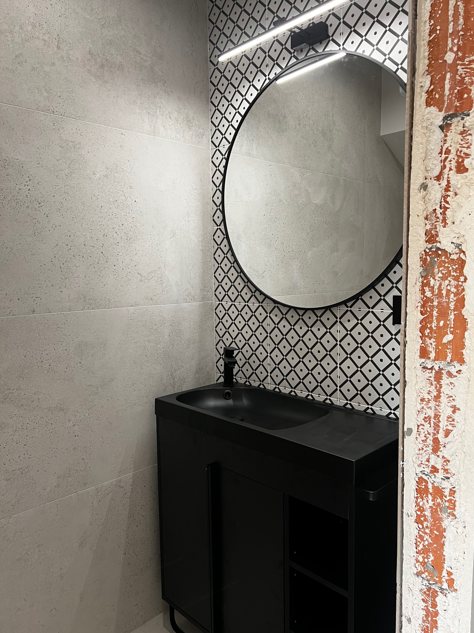Częściowo wykończona łazienka z czarną szafką pod umywalkę, okrągłym lustrem z podświetleniem LED i wzorzystymi płytkami, widoczny fragment surowej ściany.