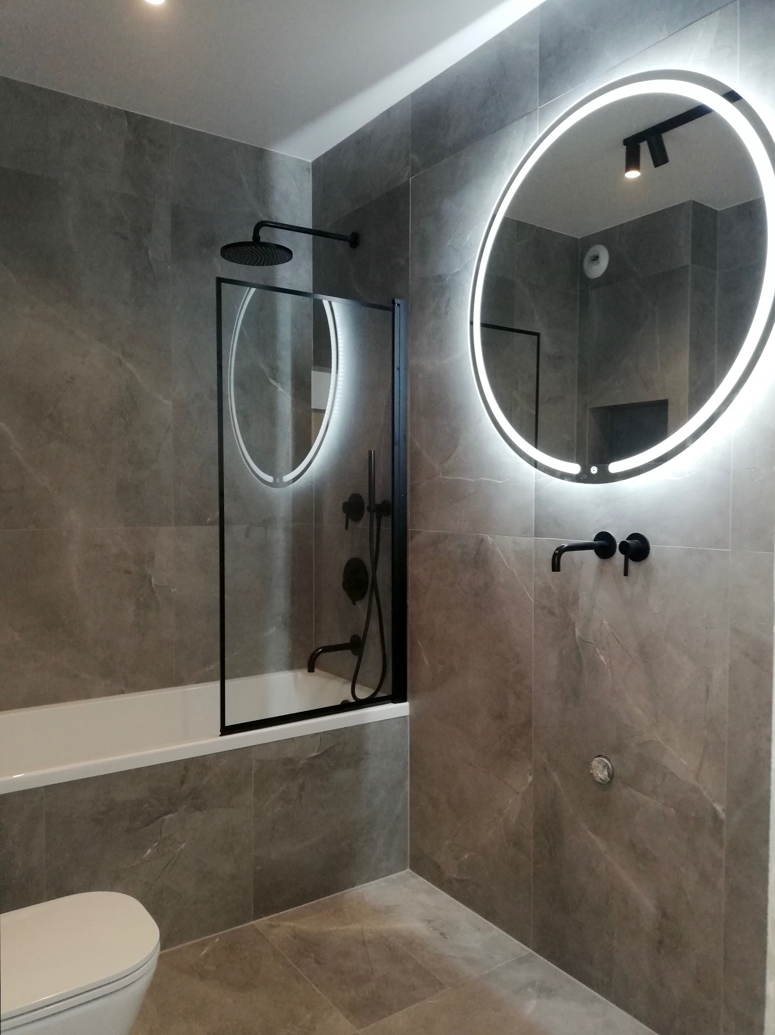Nowoczesna łazienka z szarymi płytkami imitującymi marmur, okrągłym lustrem podświetlanym LED, czarną armaturą i kabiną prysznicową z czarnymi ramami.