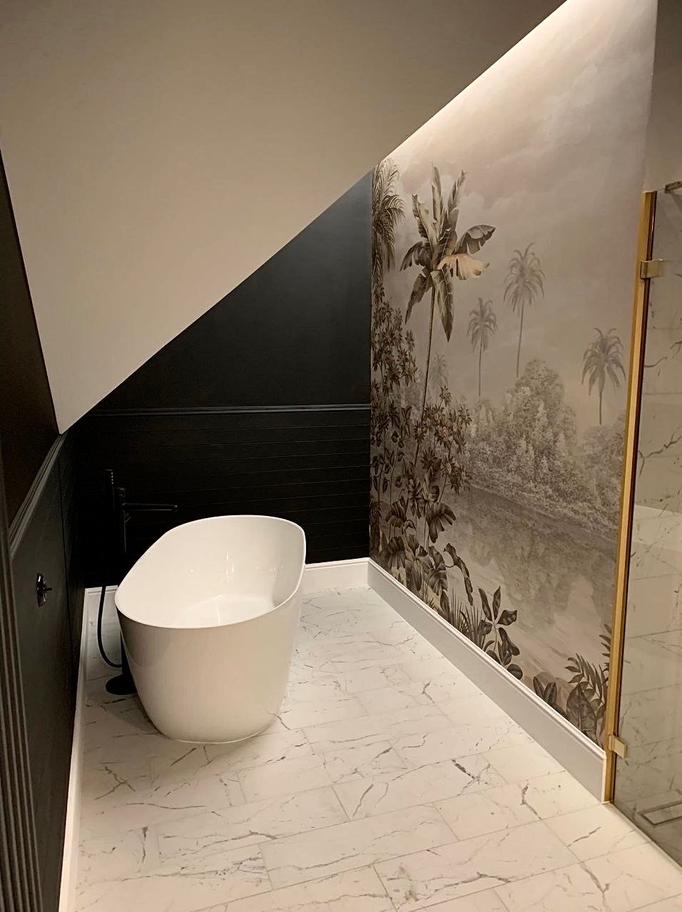 Nowoczesna łazienka na poddaszu z wolnostojącą białą wanną, marmurową podłogą i ścianą z tropikalnym motywem, oświetlona delikatnym światłem.