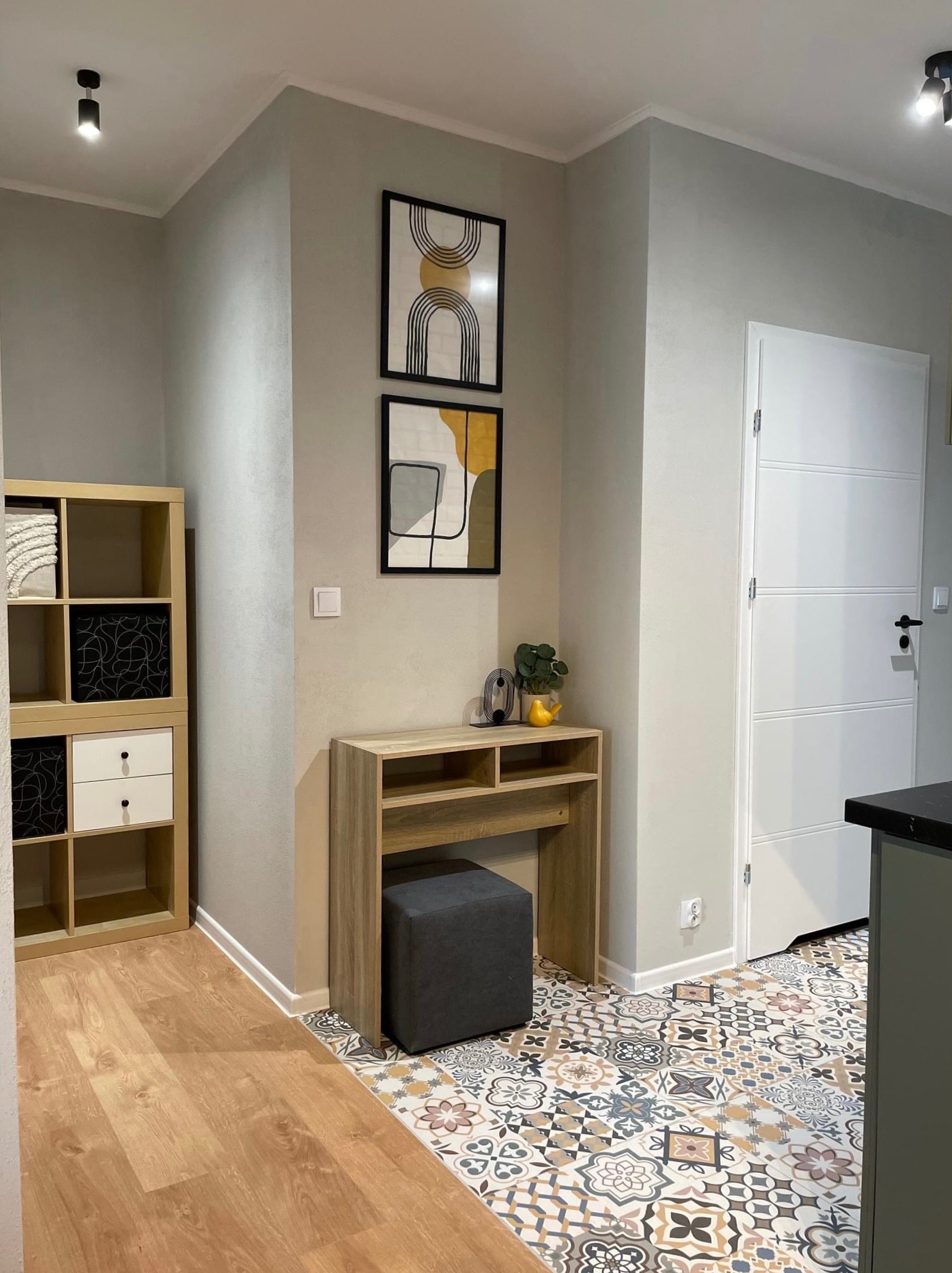 Wnętrze mieszkania z dekoracyjną podłogą w stylu patchwork, drewnianą konsolą z szarym pufem, półkami i dwoma abstrakcyjnymi obrazami na ścianie w odcieniach szarości i beżu.