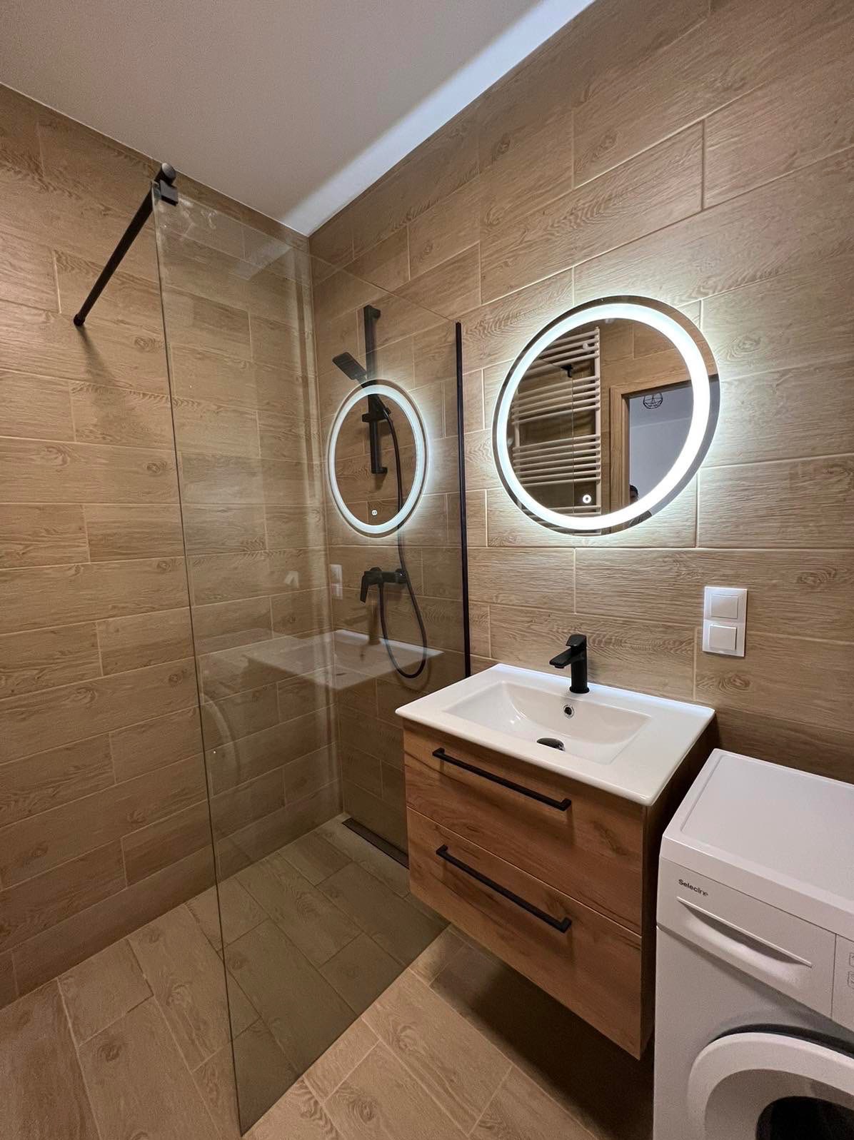 Nowoczesna łazienka z kabiną prysznicową typu walk-in, umywalką z szafką z drewnianym fornirem, dwoma okrągłymi lustrami z podświetleniem LED oraz pralką Selectric.