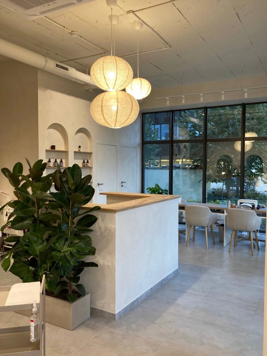 Jasne wnętrze salonu kosmetycznego z recepcją, trzema okrągłymi lampami wiszącymi z abażurami z plecionki, dużym oknem z widokiem na zieleń i rośliną doniczkową na pierwszym planie.