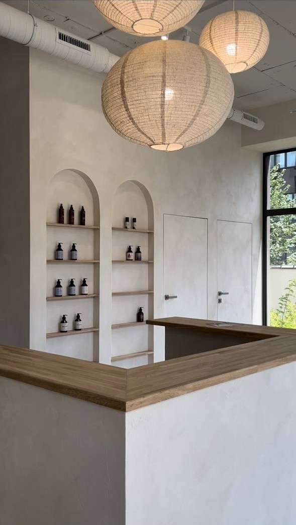 Minimalistyczne wnętrze z drewnianym blatem, wnękami z półkami na kosmetyki i trzema okrągłymi lampami z abażurami, widok na zewnątrz przez okno.