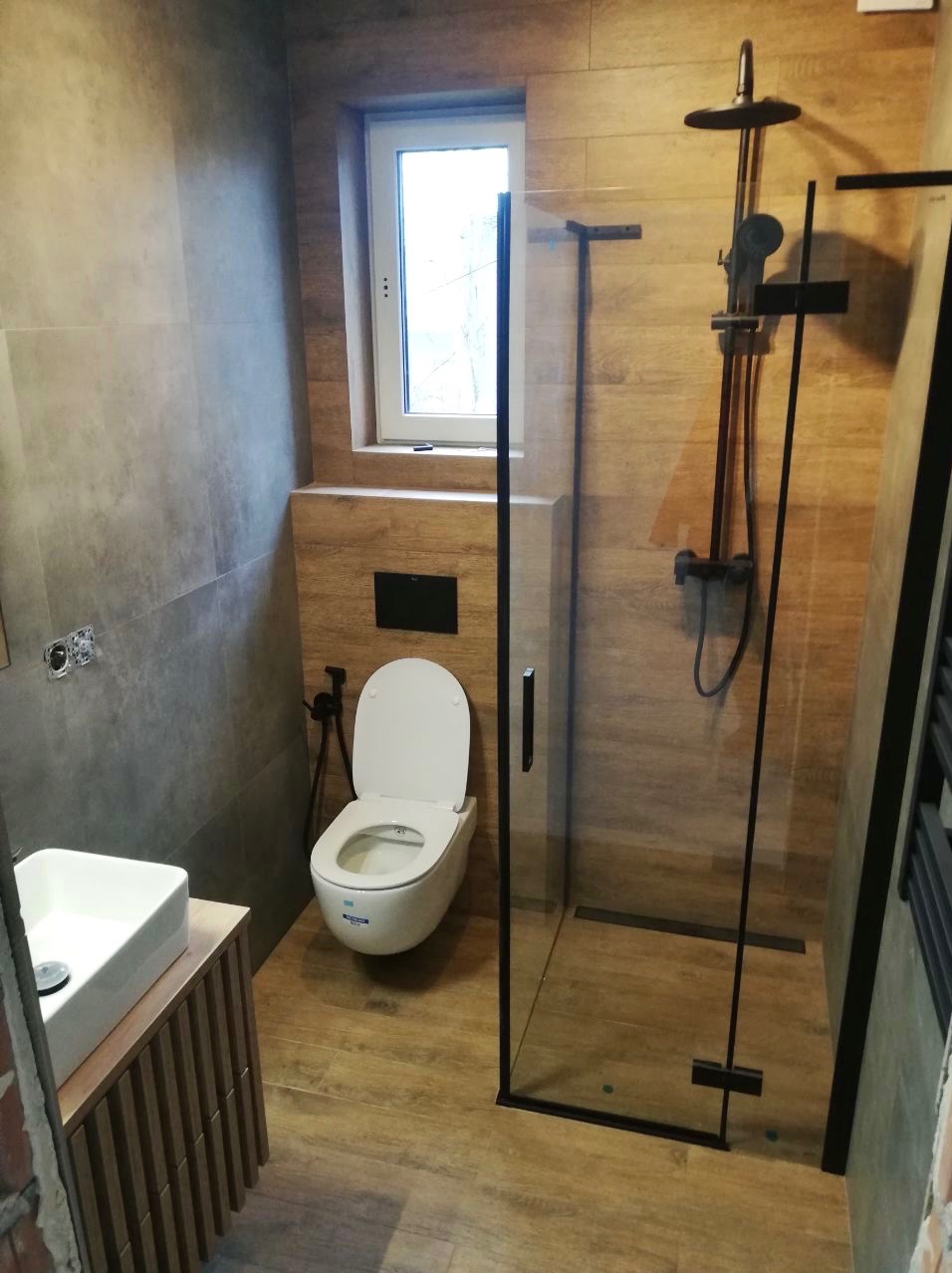 Nowoczesna łazienka z drewnopodobnymi płytkami, kabiną prysznicową typu walk-in z czarnymi profilami, podwieszaną toaletą z bidetką i umywalką nablatową.