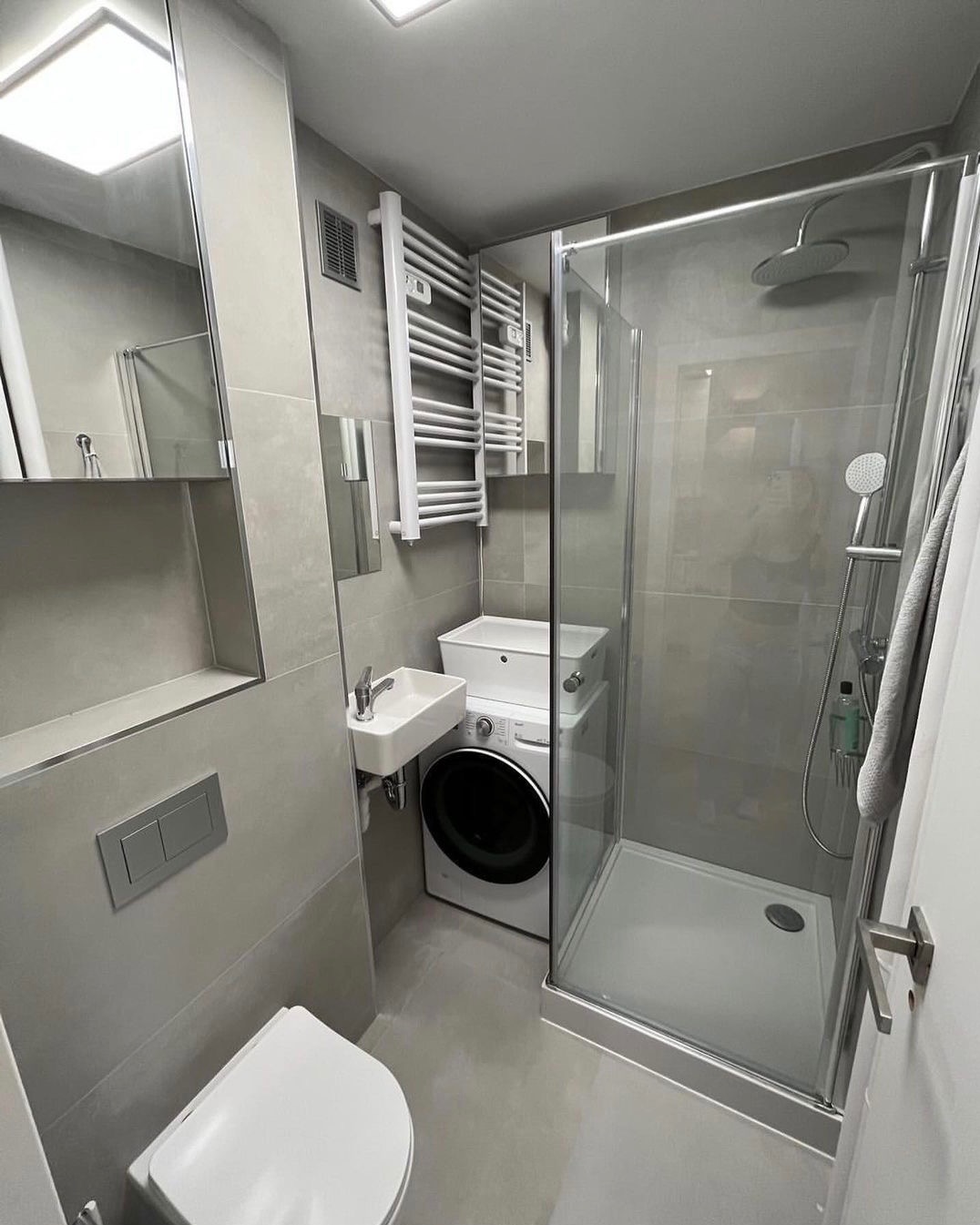Nowoczesna łazienka w odcieniach szarości z kabiną prysznicową, umywalką nad pralką, grzejnikiem drabinkowym i podwieszaną toaletą.