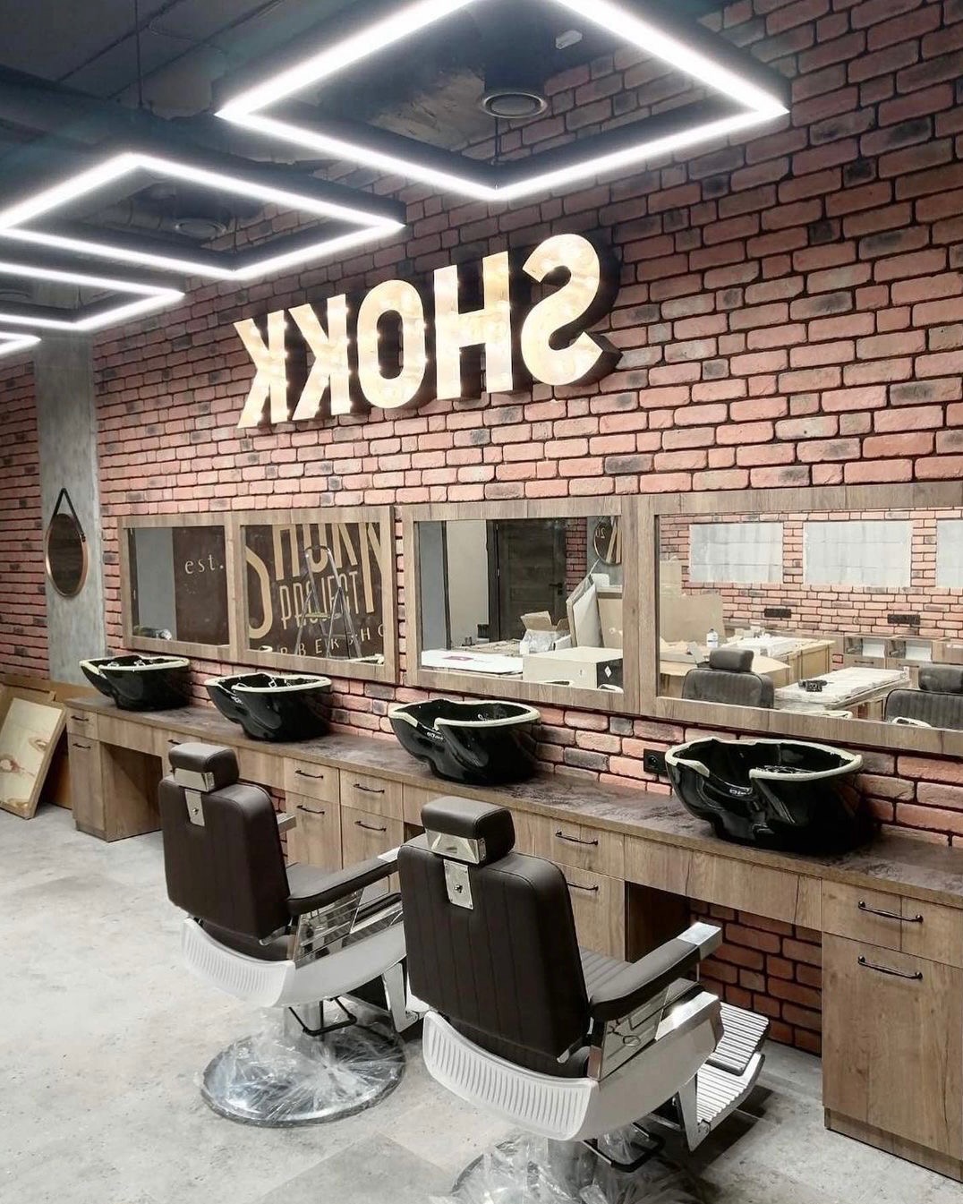 Wnętrze salonu fryzjerskiego z ceglaną ścianą, podświetlanym napisem SHOKK, czterema stanowiskami z umywalkami i lustrami oraz dwoma fotelami fryzjerskimi z brązową tapicerką i srebrną podstawą.