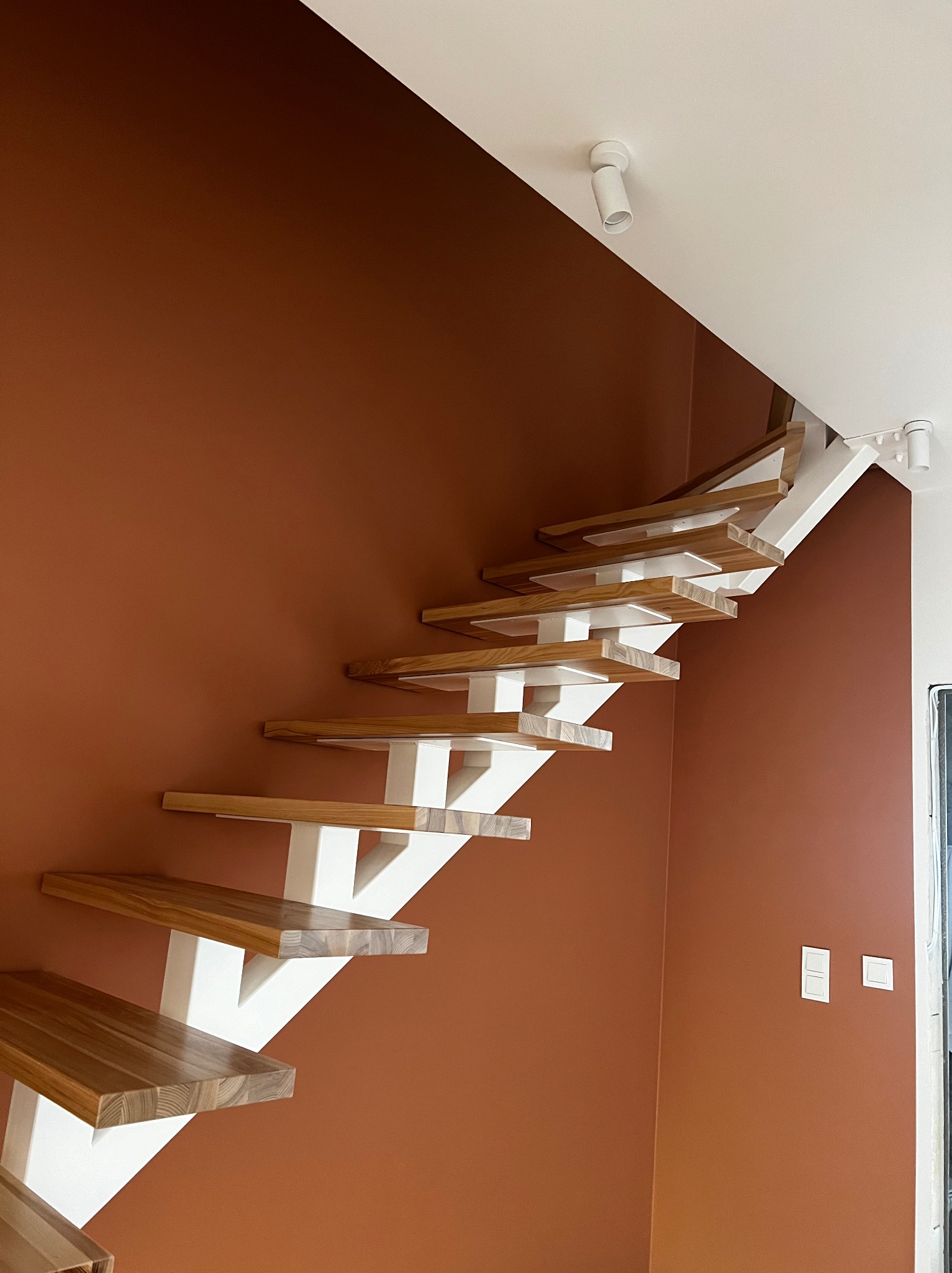 Boczne ujęcie drewnianych schodów z białymi wspornikami na tle ściany w kolorze terakoty, oświetlonych punktowymi lampami sufitowymi.
