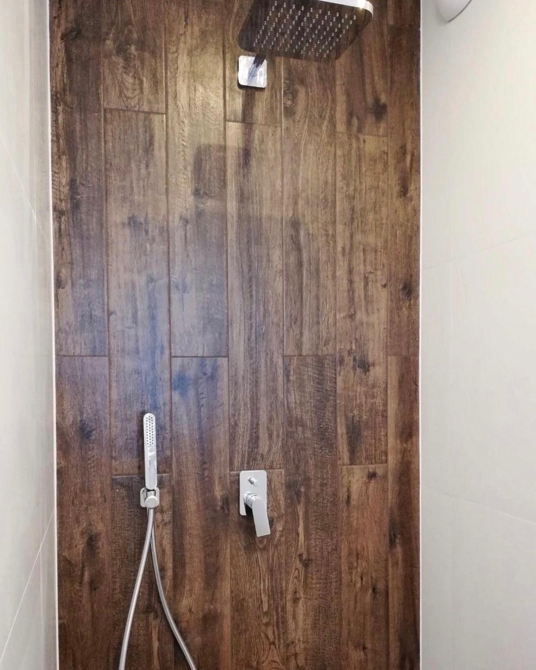 Kabina prysznicowa wyłożona płytkami imitującymi drewno, z zamontowaną deszczownicą, słuchawką prysznicową i baterią, widok z bliska.