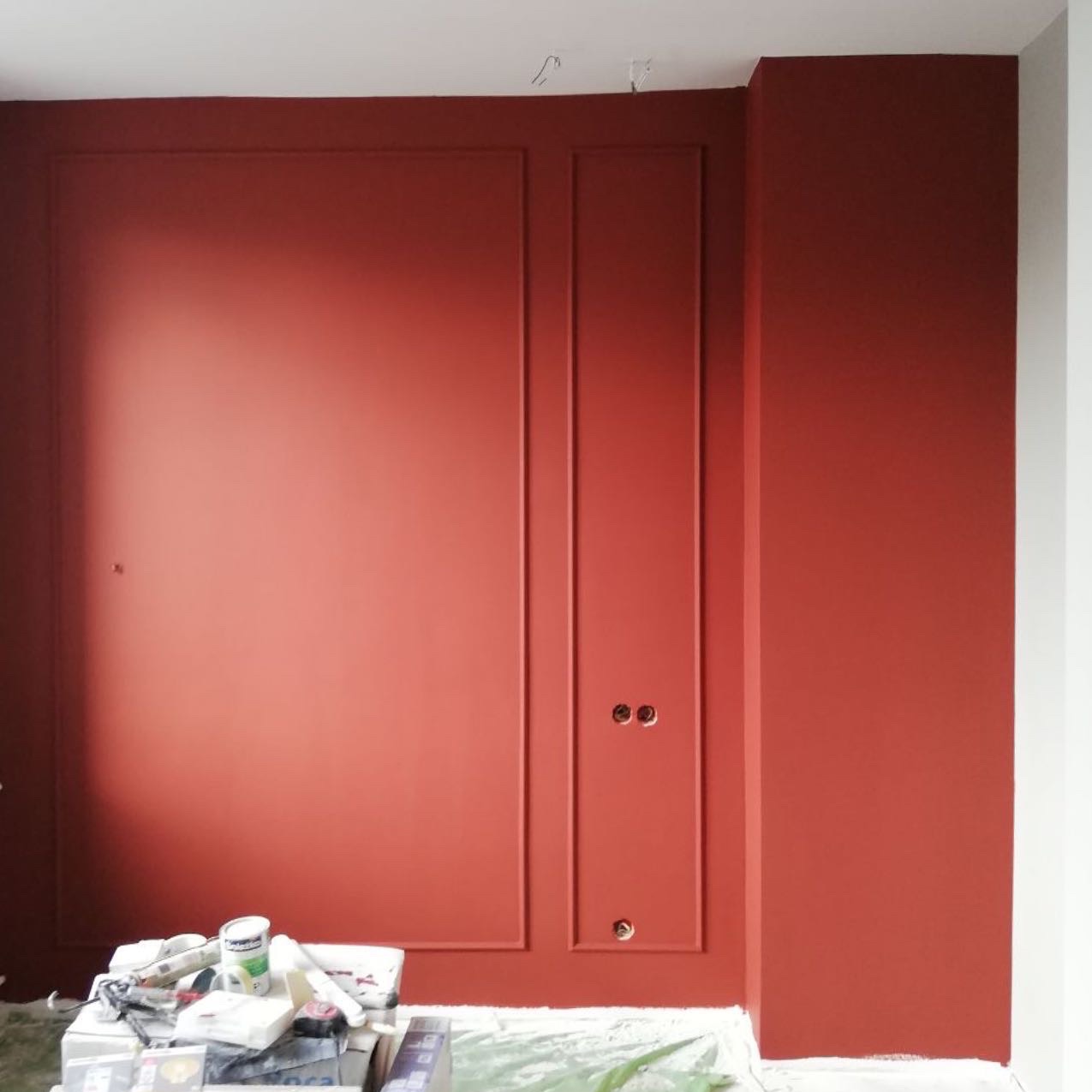 Ściana w kolorze terakoty z dekoracyjnymi listwami i przygotowanymi otworami na gniazdka, widoczne narzędzia malarskie na podłodze zabezpieczonej folią.