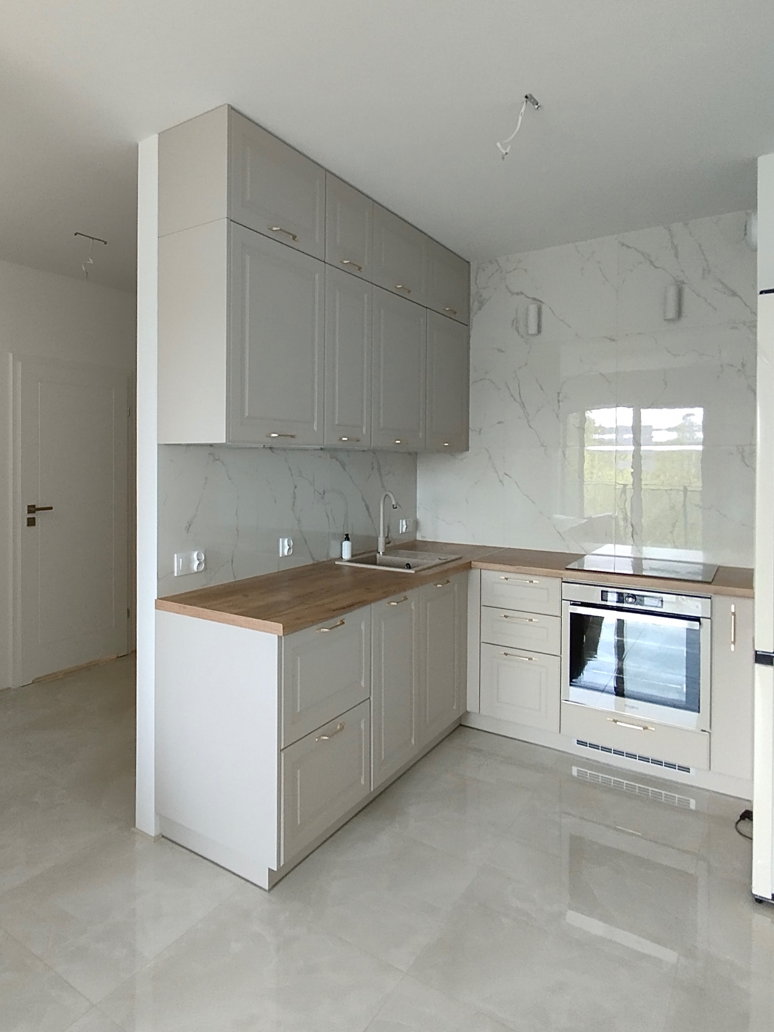 Jasna, nowocześnie urządzona kuchnia z szarymi szafkami, drewnianym blatem i marmurową ścianą, widok na okno z zielenią na zewnątrz, niedokończone oświetlenie sufitowe.
