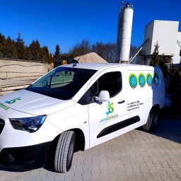 JanSerwis.pl - Biała furgonetka serwisowa z logo firmy Janserwis na tle zakładu przemysłowego z silosami i kopcami piasku, widoczny również ciężki sprzęt budowlany.